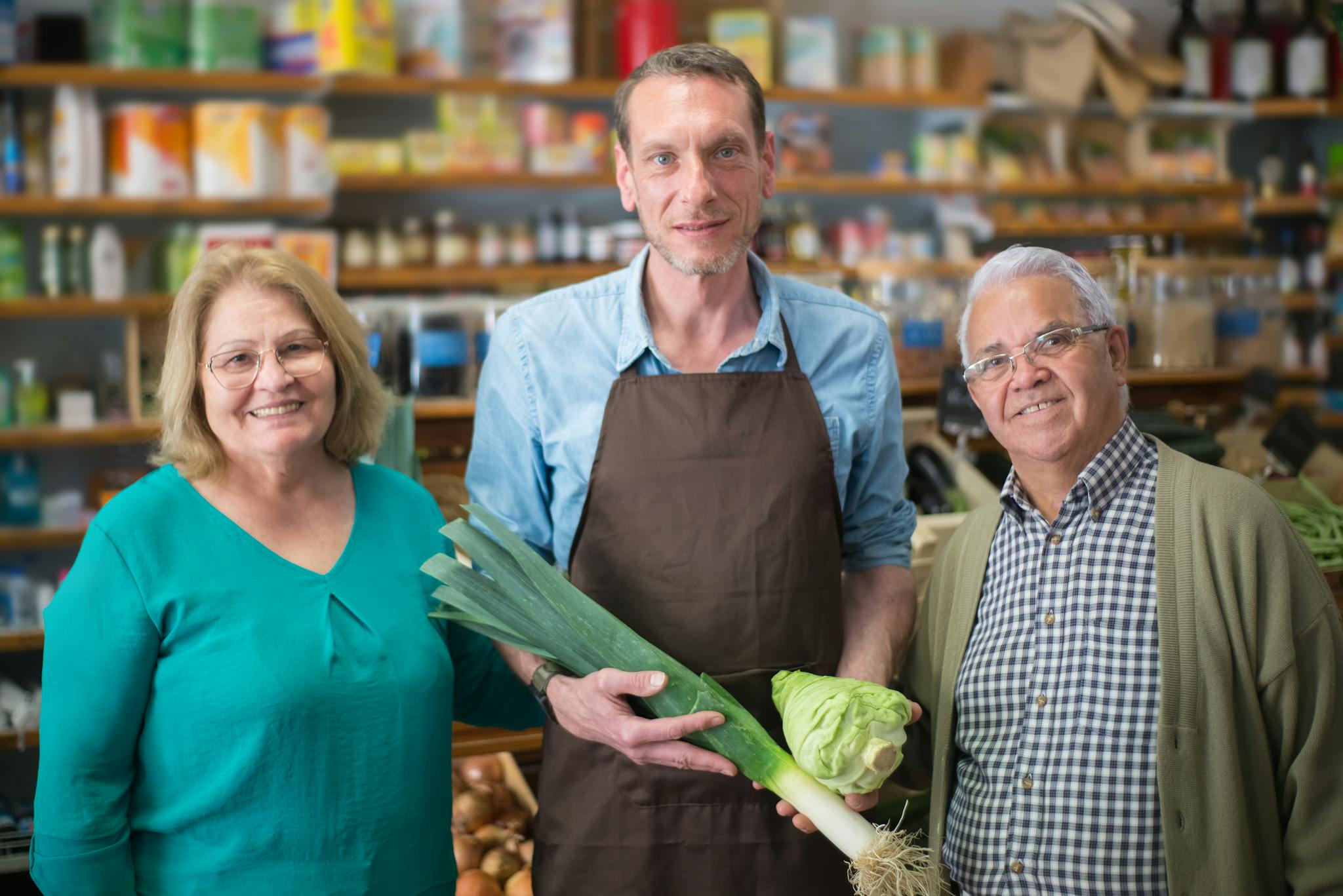 A friendly grocery store clerk presenting fresh produce with two elderly shoppers.